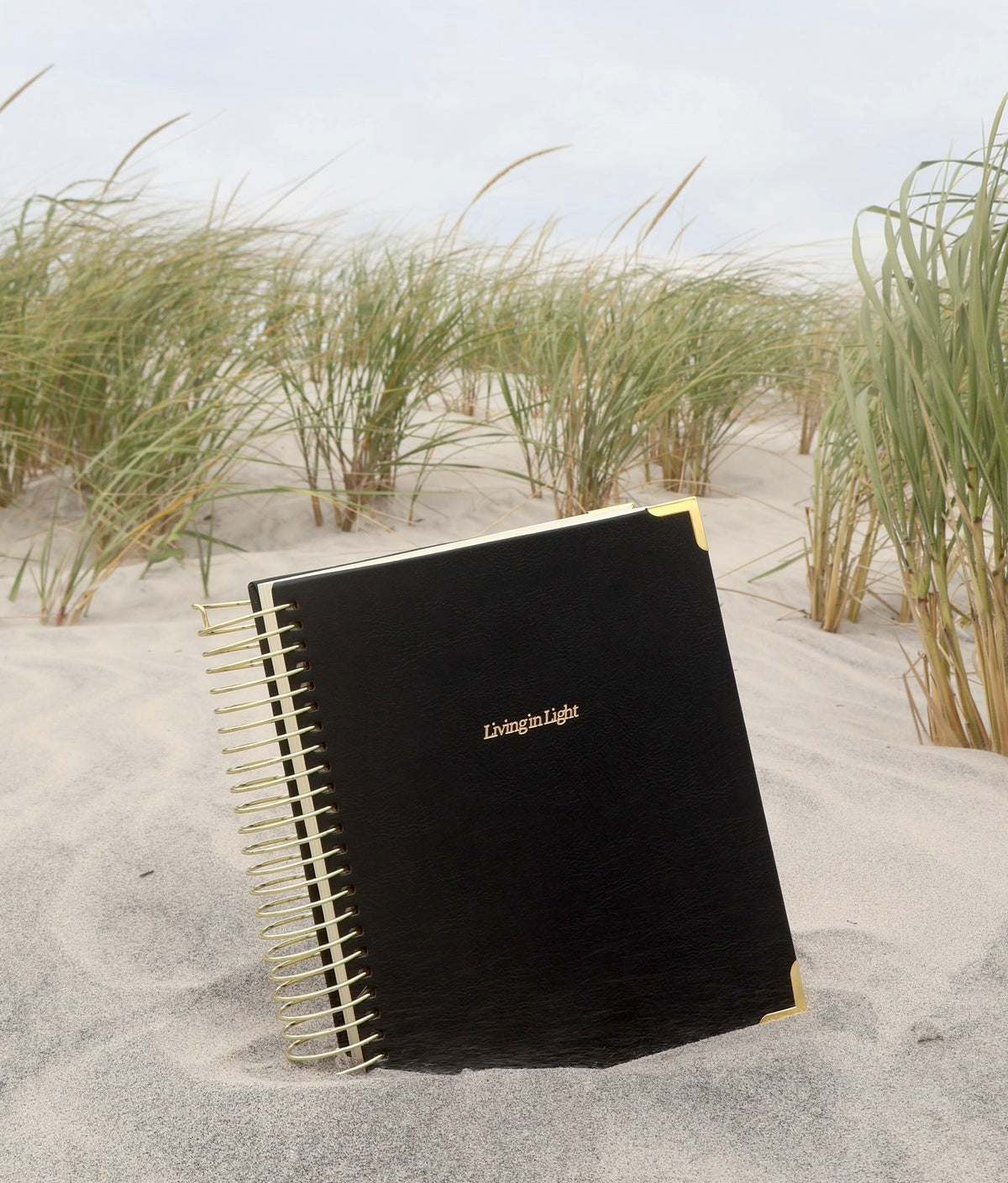 A black spiral-bound notebook with gold lettering and gold corner accents lies on sand in a beach dune setting with grasses and a cloudy sky in the background.