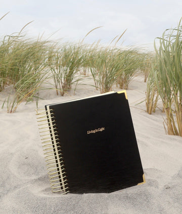 A black spiral-bound notebook with gold lettering and gold corner accents lies on sand in a beach dune setting with grasses and a cloudy sky in the background.